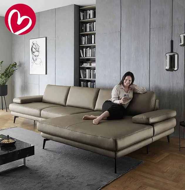 Modernes Wohnzimmer mit großem olivfarbenem Ledersofa und breiter Recamiere auf schwarzen Metallfüßen. Frau sitzt entspannt mit Smartphone, graue Wand mit Bücherregal und Bild, dunkler Teppich und eleganter Couchtisch im Vordergrund.